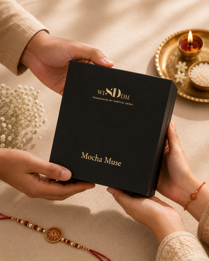 A black rectangular gift box with gold lettering reading 'WISDOM Fragrances by Sheetal Desai' and 'Mocha Muse' is held by two hands over a beige fabric surface. Decorative elements include a gold tray with a lit candle, white flowers, and a red-and-gold Rakhi.