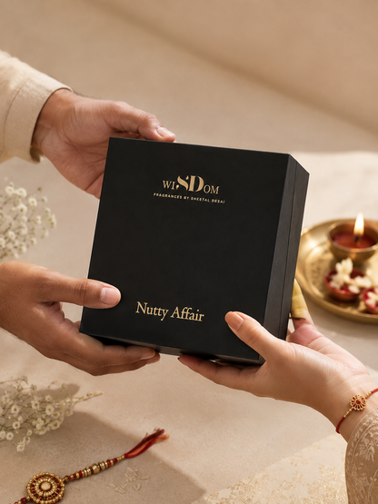 A black gift box with gold lettering reading 'wisdom' and 'Nutty Affair' is being held by multiple hands in a warm, festive setting with a lit diya in the background. Rakhi special collection