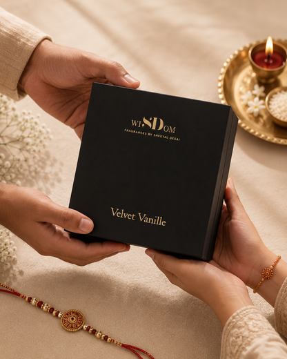 A black rectangular gift box with gold lettering reading 'WISDOM Fragrance of Sacred Heart' and 'Velvet Vanille' is held by two hands over a beige fabric surface. A gold tray with a lit candle and a red-and-gold Rakhi are visible in the background and foreground.