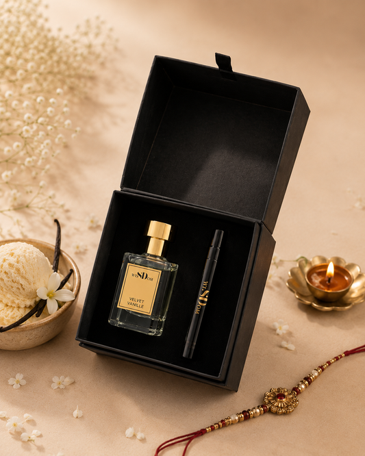 A black gift box opened to reveal a clear perfume bottle named Velvet Vanille with a gold cap and a black cosmetic stick with gold lettering, styled on a beige surface with dried flowers, a small bowl, and a lit gold candle.